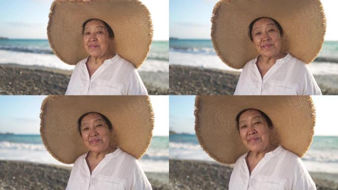 戴着草帽的快乐亚洲老妇人看着相机微笑着在风景如画的地中海海岸摆姿势，背景是波浪在慢动作中滚动。积极享