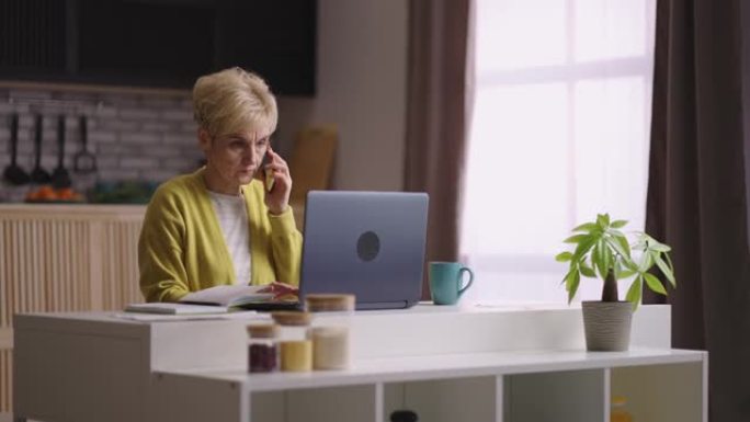 老年妇女在家工作，坐在厨房里用手机打电话，用笔记本电脑，中等肖像