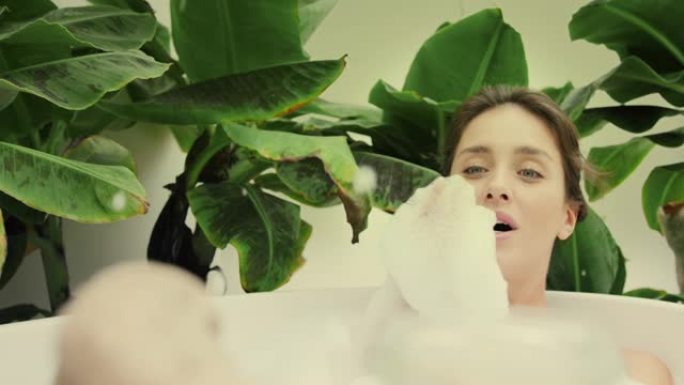 年轻女子在浴室内郁郁葱葱的绿色花园里洗泡泡浴。吹出泡沫