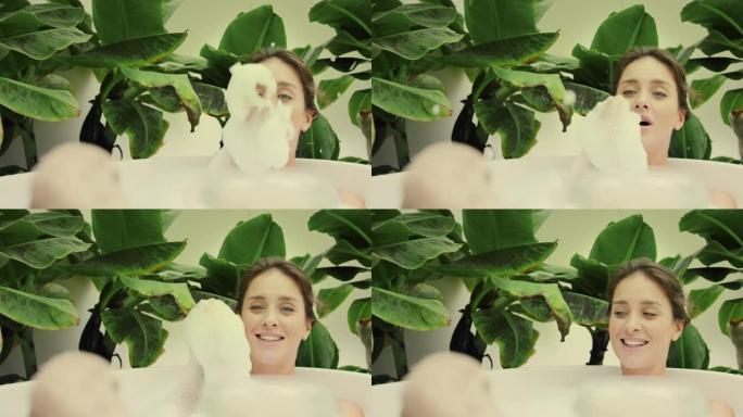 年轻女子在浴室内郁郁葱葱的绿色花园里洗泡泡浴。吹出泡沫