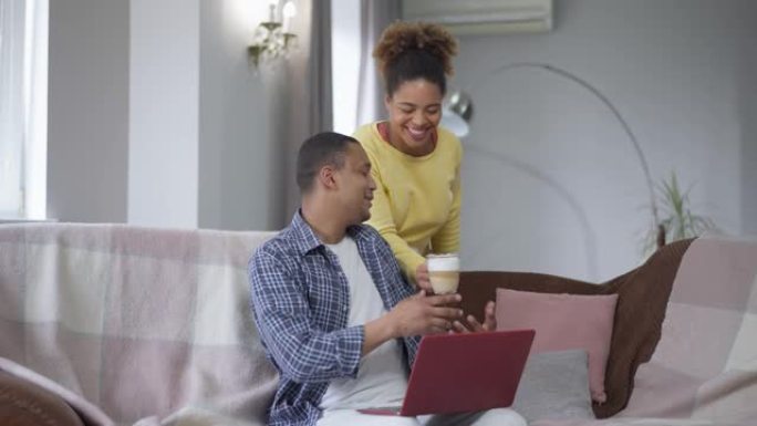 充满爱心的非裔美国妻子为忙碌的丈夫在家庭办公室在线发送消息带来咖啡杯。微笑美丽的女人照顾过度劳累的男