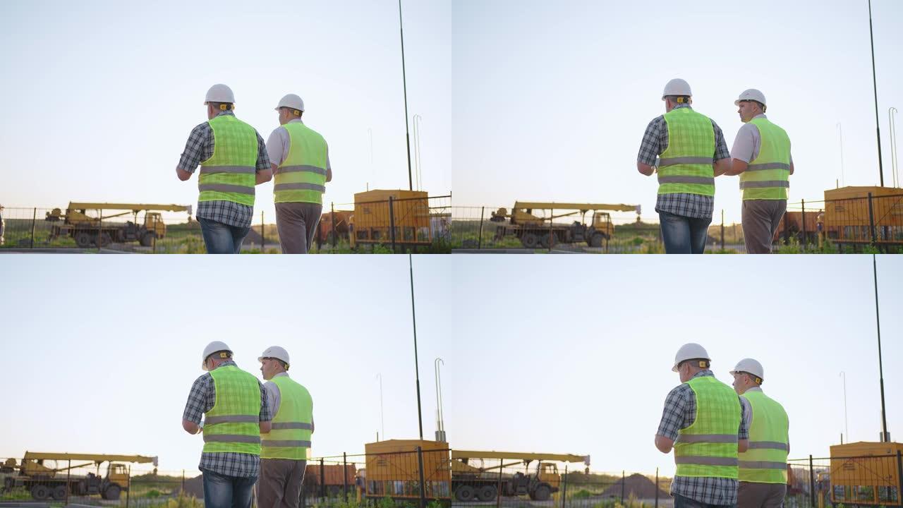两名穿着安全服的中年男性建筑工人的腰部站立在施工现场，男子使用对讲机，他的同事拿着带有项目计划的纸