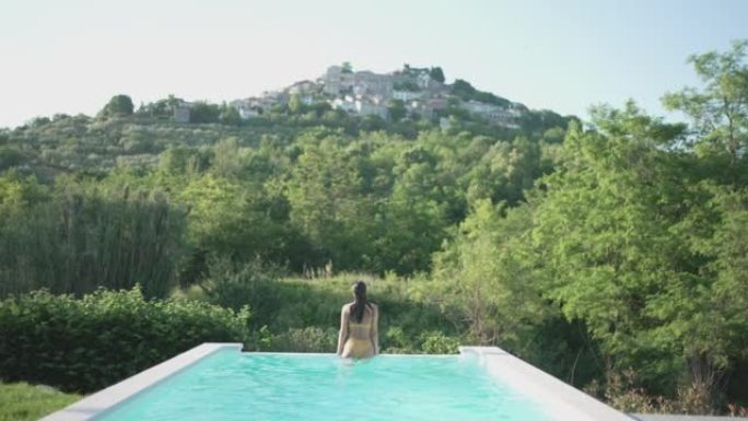 年轻女子在大自然的游泳池中游泳，俯瞰一个小村庄