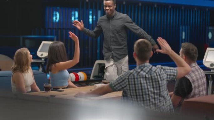 一个快乐的非洲裔美国人跑到朋友面前，把五个不同种族的人放在一个保龄球俱乐部的桌子旁。为了庆祝胜利，祝