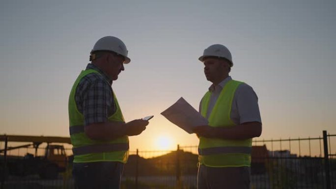 施工经理和工人在施工现场握手。带着平板电脑的建筑工人和一名戴着白盔的男子检查员在日落时站着握手。协议