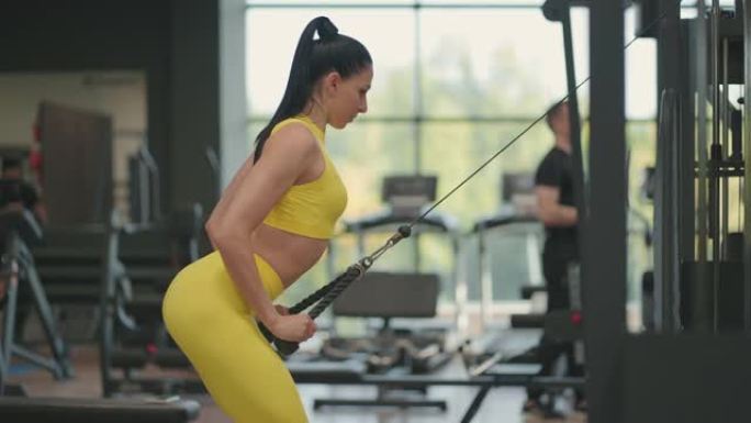 一位穿着黄色运动服的年轻西班牙裔妇女在交叉运动中进行锻炼，从上方拉一根绳子训练背部和肩膀。黑发女人在