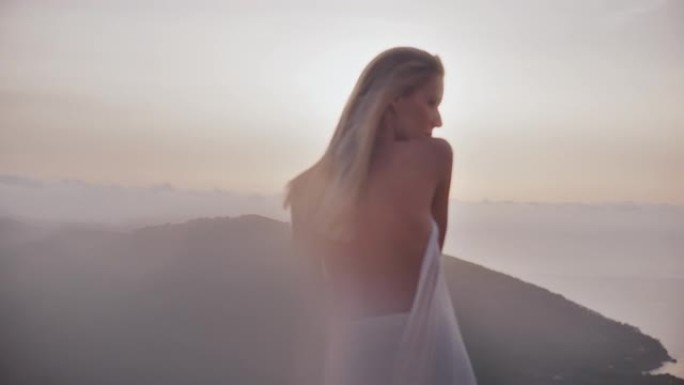 美丽的天真无邪的云朵女神，金色的头发裹着白色的长袍，顶着夕阳站在山顶上，用相机调情