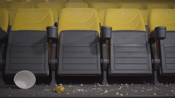 摄像机沿着黄色椅子的肮脏空电影院移动。电影院，地板上有爆米花和香蕉皮。参观者之后的电影院。