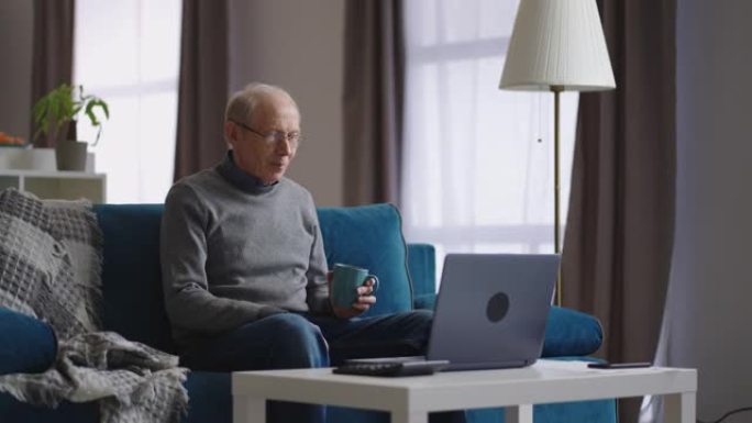 男性退休人员正在使用笔记本电脑通过与朋友或家人的视频聊天，看着网络摄像头和喝茶进行交流