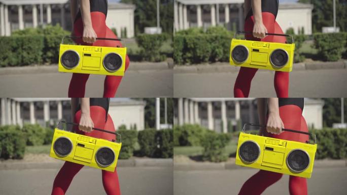 穿着红色腿的面目全非的苗条女人穿着老式黄色录音机走在阳光明媚的夏日街上。适合白人20世纪80年代女运