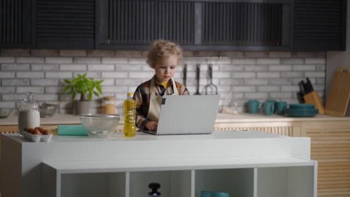 有趣的小男孩穿着像厨师在现代公寓的厨房里玩耍，用笔记本电脑准备做饭