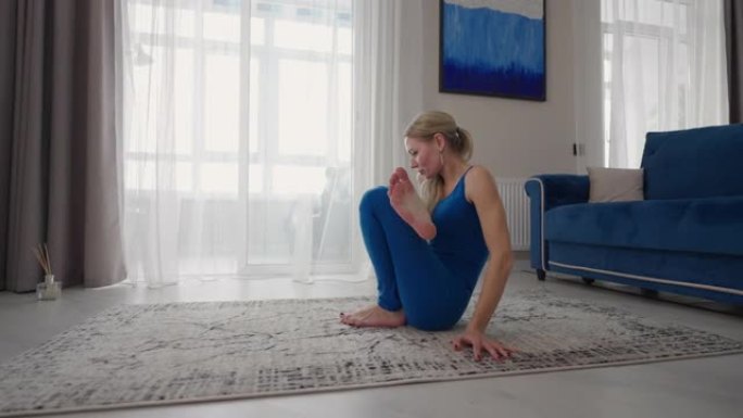 在家的女人足不出户就做瑜伽练习。家庭培训。一个女人穿着蓝色西装在地毯上训练
