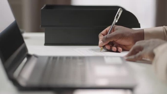 非裔美国人正在纸上写笔记，坐在笔记本电脑的桌子上，在线学习，双手特写