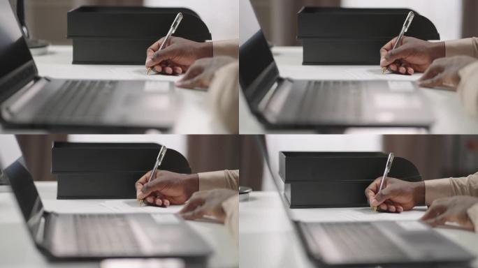 非裔美国人正在纸上写笔记，坐在笔记本电脑的桌子上，在线学习，双手特写