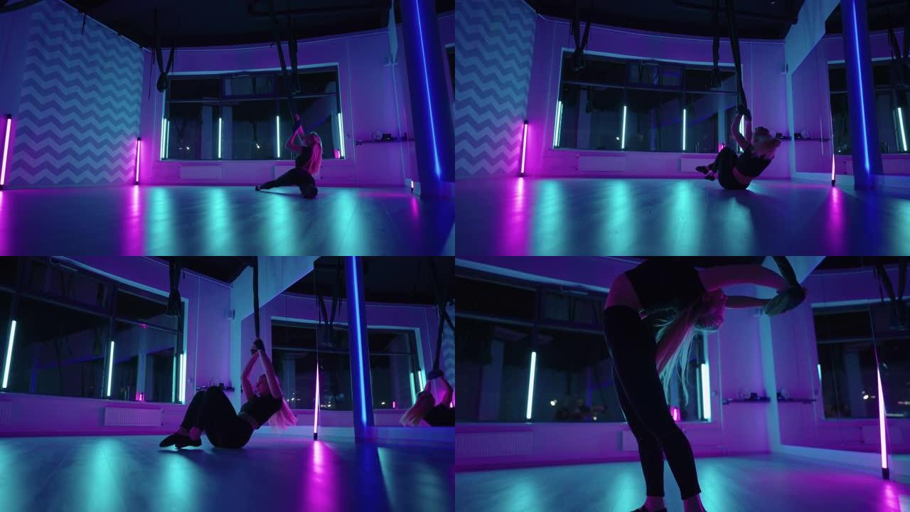 一个女人在霓虹灯下做瑜伽并在悬吊的吊床上伸展。年轻漂亮苗条的身体健身女孩在健身房练习飞行瑜伽。