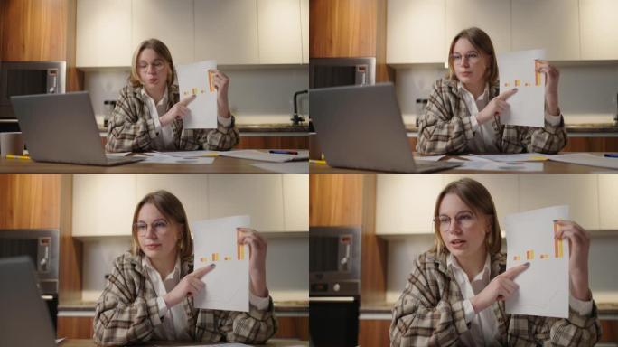 坐在家里的厨房里，戴着眼镜的女人在笔记本电脑的相机上显示了一张图表。家庭办公室远程工作。用图表和表格