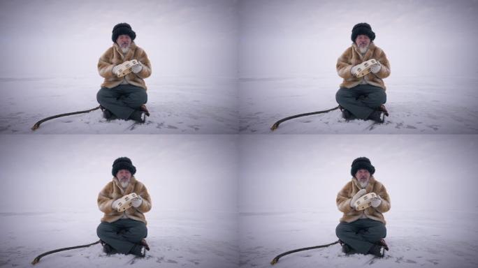 宽镜头老蒙古人坐在冰冻的雪地上，在户外慢动作玩手鼓。背景下具有北极极地性质的集中自信的高级男子的前视