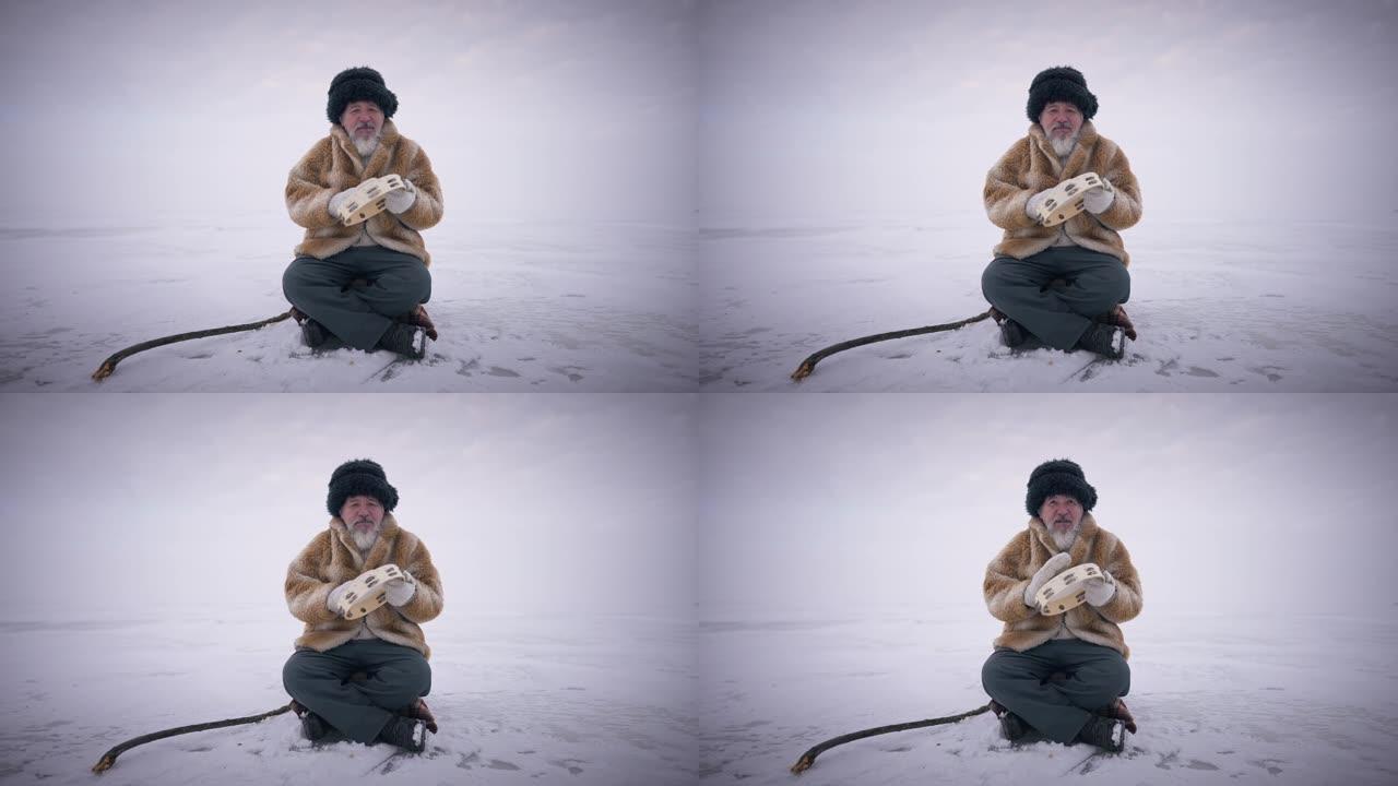 宽镜头老蒙古人坐在冰冻的雪地上，在户外慢动作玩手鼓。背景下具有北极极地性质的集中自信的高级男子的前视