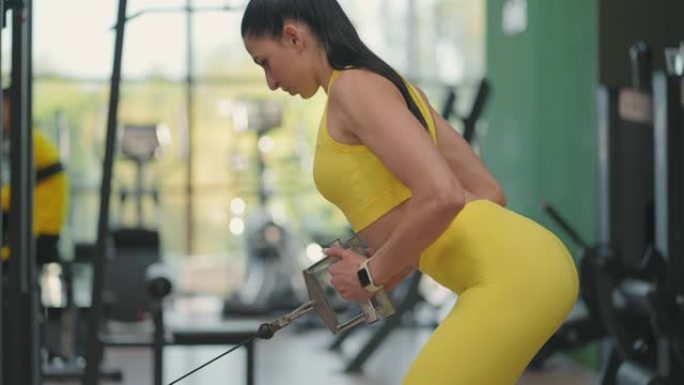 一位穿着黄色运动服的年轻西班牙裔妇女在跨界运动中进行锻炼，从下方拉出一根钢绳来训练她的背部。一个女人
