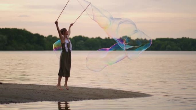 一位年轻的女艺术家展示了一个肥皂泡表演，在日落时在湖岸上吹起巨大的肥皂泡。在慢动作中展示美丽的肥皂泡