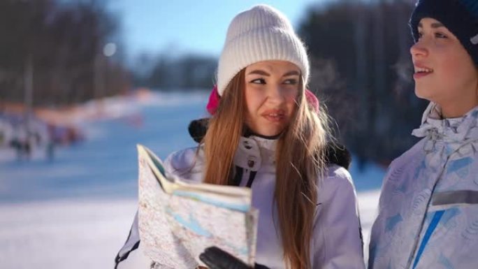 两名年轻妇女使用纸质地图选择路线，离开阳光明媚的滑雪胜地。自信的女性高加索朋友在户外聊天的肖像。