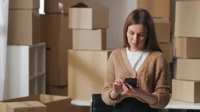 年轻女子是小企业的老板，站在公寓的仓库里，用智能手机查看商品的照片