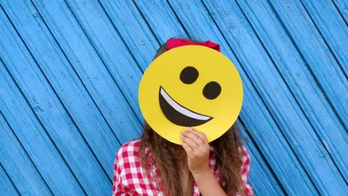 一件格子衬衫，头发上有一个红色蝴蝶结。女孩在纸板脸后面藏了一张快乐的笑脸。世界表情符号日。做自己的幸