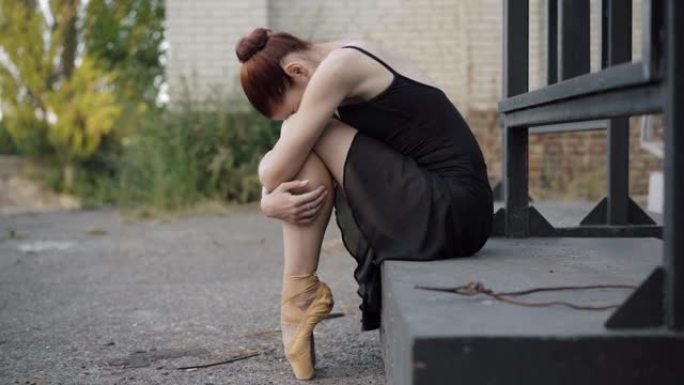 沮丧的苗条芭蕾舞演员坐在户外拥抱膝盖的侧视图。悲伤的年轻白人妇女，穿着黄色尖，脚尖和黑色连衣裙。沮丧