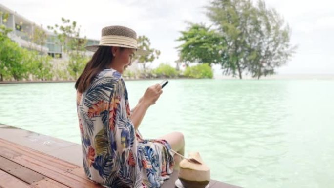 女人坐在游泳池边喝椰子水放松玩手机。