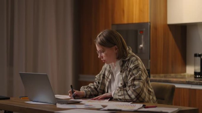 戴眼镜的女人在家庭办公室里远程工作，桌子上放着笔记本电脑，并在图表上记录数据，商业经济学家分析师