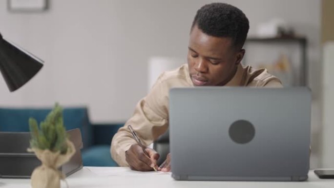 美国黑人学生在公寓里工作，在纸上写作，坐在笔记本电脑的桌子旁，在线学习