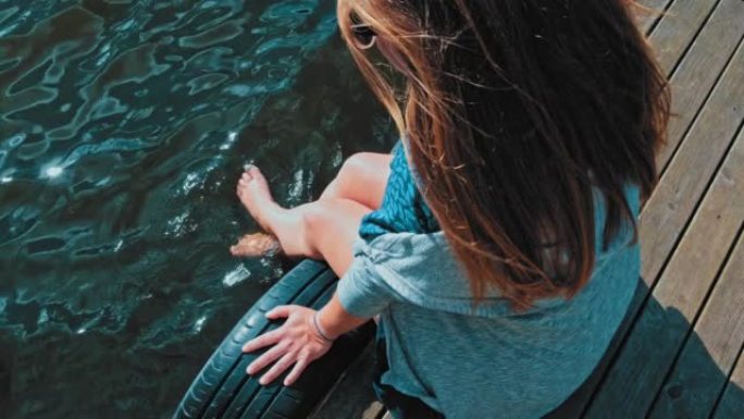年轻的白人妇女脚踢水坐在边缘的木制码头码头上，看着平静的湖港