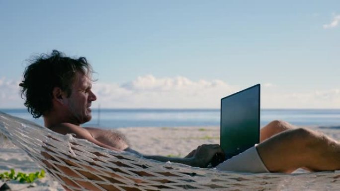 男子用笔记本电脑躺在海滩上两棵棕榈树之间的吊床上