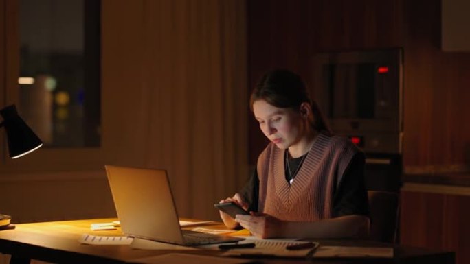 美丽的女人在家工作到很晚，使用笔记本电脑并在智能手机上接收短信。女人的手在手机上发短信，用于在社交网
