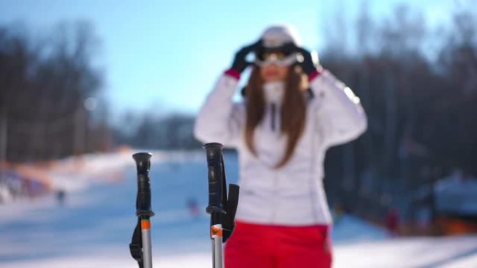 冬季度假胜地阳光直射的滑雪杖，模糊的高加索年轻女子从背景中缓慢行走，戴上滑雪护目镜。快乐自信的健康女