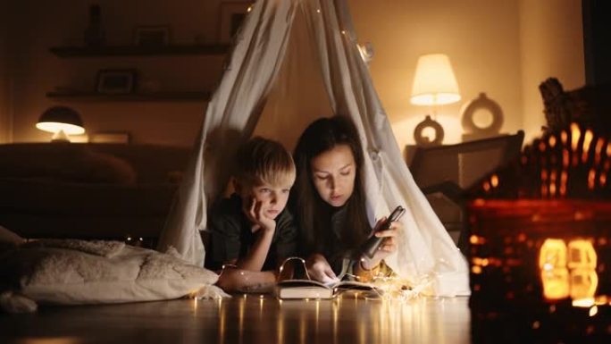 西班牙裔母亲和儿子在灯笼的灯光下一起读书，在黑暗中在家中的帐篷在花环和灯的灯光下一起读书。家庭和家庭