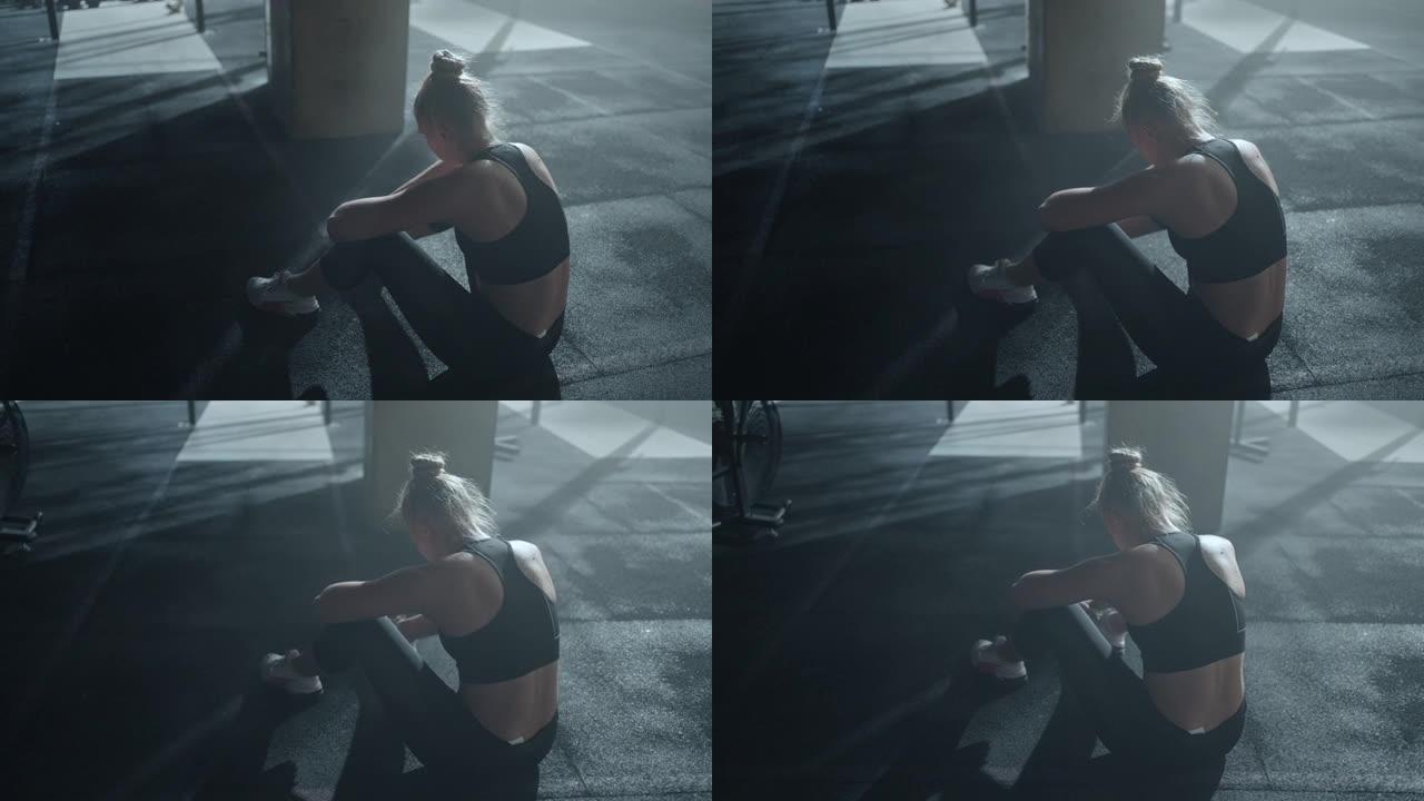 疲惫的女运动员在健身房辛苦训练后哭泣，独自坐在地板上，遭受身体的疼痛