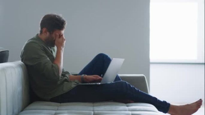 疲惫的男人正在家里用笔记本电脑工作，坐在沙发上打字，感到疼痛和疲倦