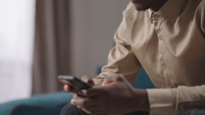 美国黑人正在坐在公寓客厅的智能手机上上网，在社交网络上查看照片