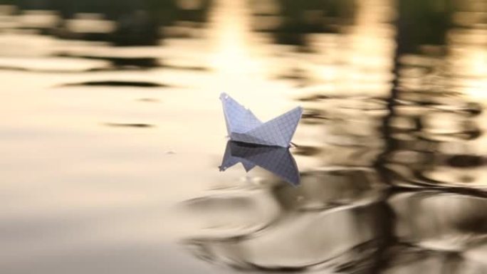 一艘纸船在美丽的日落时漂浮在水中的波浪上。折纸船航行。梦想，未来，童年，自由或希望的概念。慢动作。高