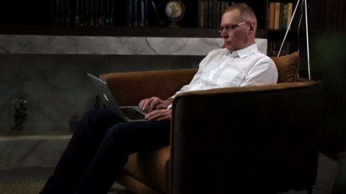 明斯克白俄罗斯2022年4月3日成熟的商人晚上工作，笔记本电脑放在腿上，坐在黑暗房间的扶手椅上