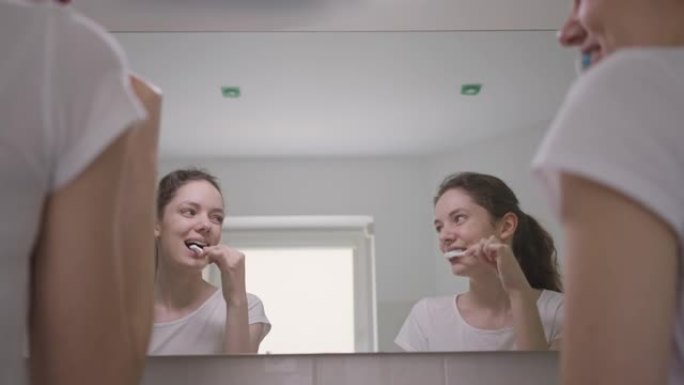两个快乐的双胞胎早上在浴室里笑着刷牙的镜子里的反射。在家拍摄苗条年轻美丽的高加索姐妹的肩膀。常规和美
