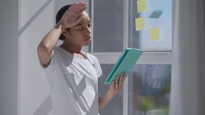 悲伤疲惫的担心的非洲裔美国妇女擦着额头，看着站在窗户上的阳光中的平板电脑屏幕。在家庭办公室等待电子邮