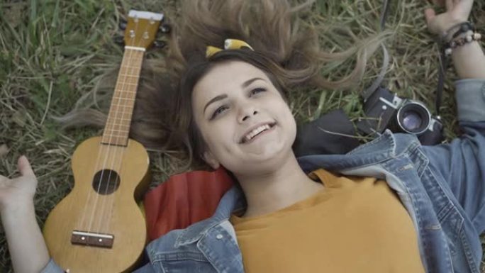 迷人的无忧无虑的高加索嬉皮士女人躺在草地上，睁开眼睛，对着镜头微笑。快乐美丽的女士在20世纪60年代