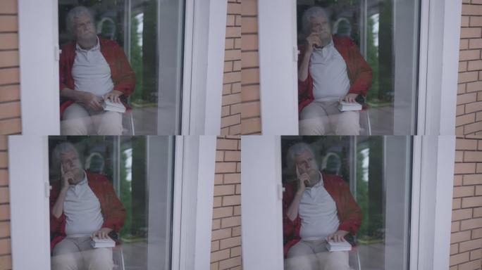 沮丧的体贴的高级残疾人合上书，看着窗外的玻璃门。室内沮丧孤独的高加索大胡子残疾退休人员的肖像。慢动作