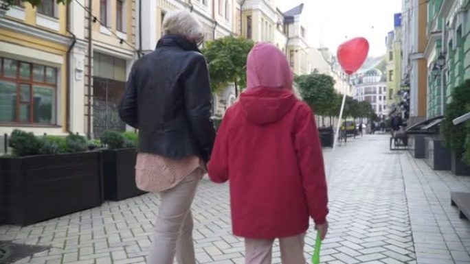 后视图白人祖母戴着冠状病毒面具与孙女在城市街道上交谈。快乐的奶奶和女孩在户外散步，享受新型冠状病毒肺