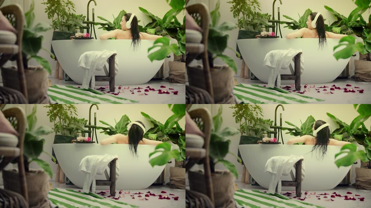 美丽的女人在现代浴室放松。在豪华的带有热带植物的浴室里听音乐