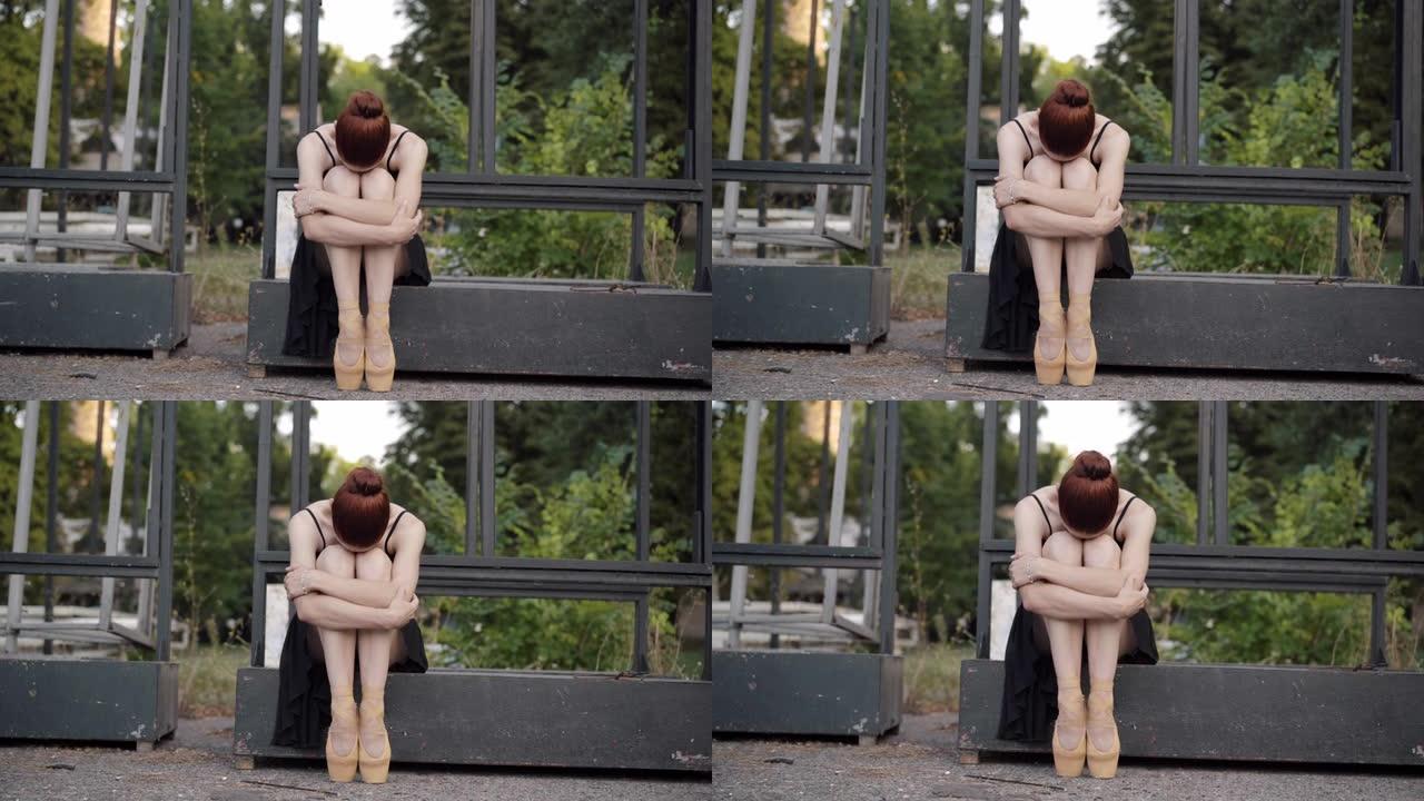 沮丧悲伤的年轻芭蕾舞演员坐着拥抱膝盖的前视图。沮丧的白人红发女人穿着脚尖鞋，穿着厌倦了舞蹈艺术。抑郁