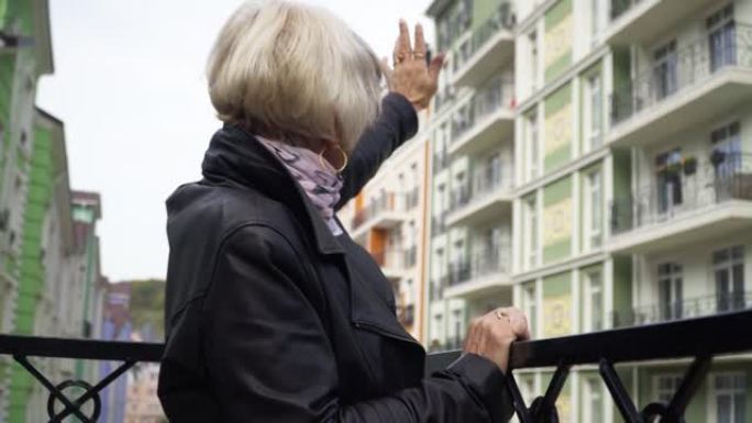 侧视图高级妇女向站在城市城市阳台上的邻居挥手。积极的高加索女性退休人员问候朋友微笑打手势。生活方式概