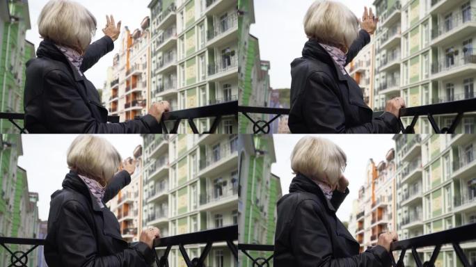 侧视图高级妇女向站在城市城市阳台上的邻居挥手。积极的高加索女性退休人员问候朋友微笑打手势。生活方式概